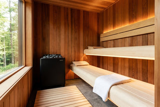 Home sauna with warm Western Red Cedar walls and ceiling, pale clear-grade aspen benches with LED glow underneath, black electric heater, and a removable wooden duckboard on a stone tile floor