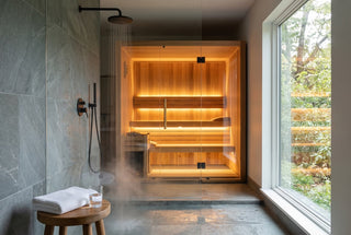 Home spa with a modern walk-in rain shower beside a glowing cedar sauna with LED bench lighting, a wooden stool with a folded white towel and a glass of water in the foreground