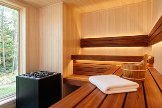 Home sauna with pale hemlock wall and ceiling lining and reddish western red cedar benches, warm LED glow under the benches, black electric heater, folded white towel and wooden bucket