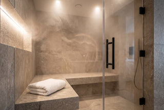 Interior of a modern premium steam room with neutral stone tile walls and bench, a glass door with matte black hardware, soft warm lighting, gentle wisps of steam, a folded white towel