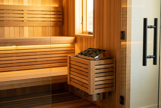 Wall-mounted electric sauna heater with stones in a cedar home sauna, glass door with matte-black hardware