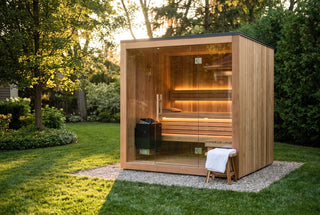 Freestanding cedar sauna pod with a glass front on a gravel pad in a green backyard at golden hour, warm-lit benches and a matte-black heater inside, a white towel on a wooden stool beside it