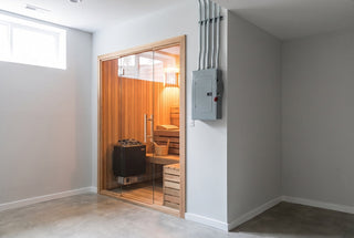 Basement home sauna with a glass door and an electric heater with stones inside, next to a gray electrical box with a disconnect handle and metal conduit mounted on the wall outside the sauna